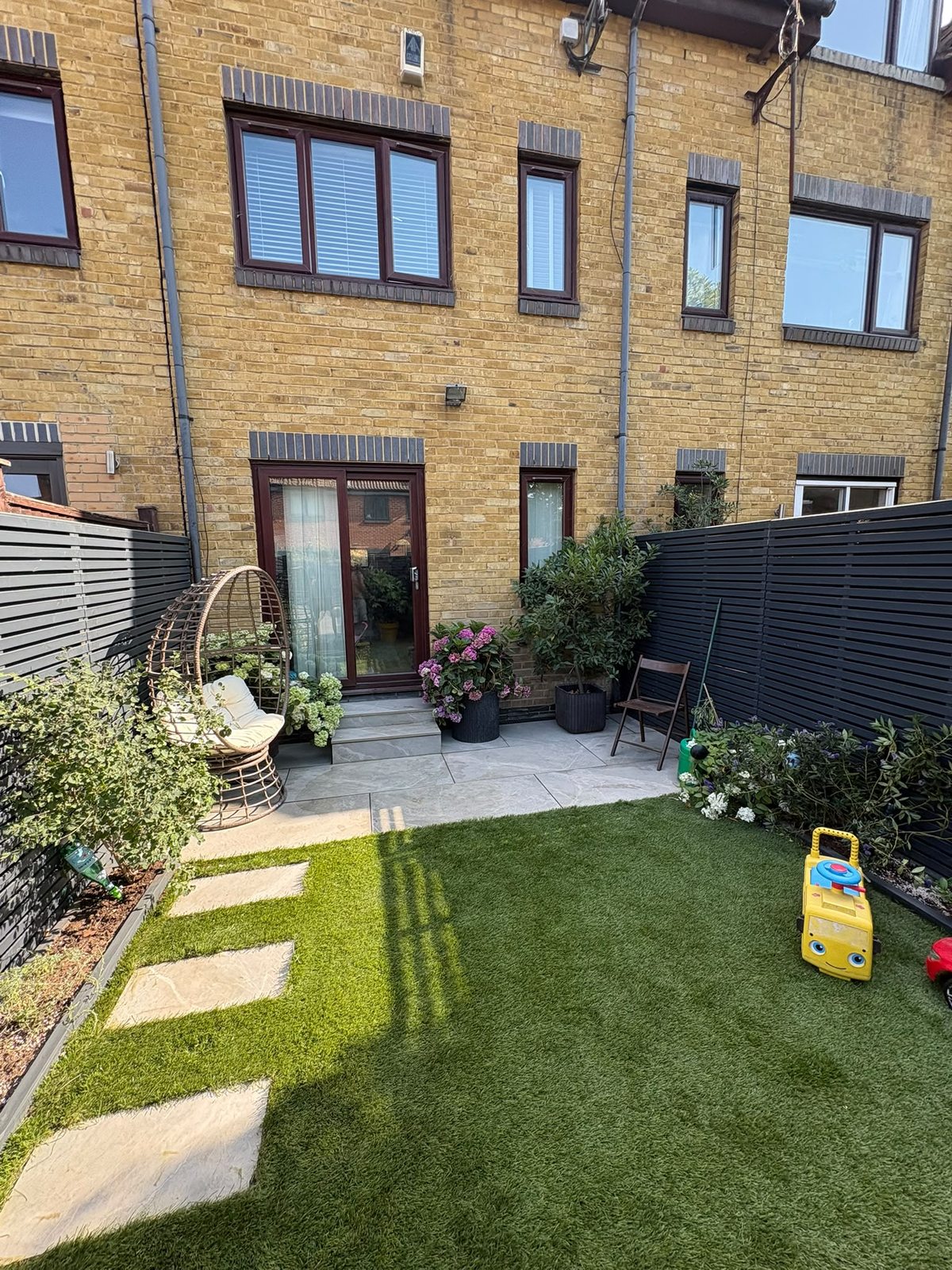 Stone patio with steps, planting and artificial lawn against black slatted fencing, Rotherhithe SE16